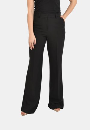 Donna con pantaloni neri a vita alta e gamba larga con piega frontale, mani nelle tasche, abbinati a décolleté beige open toe su sfondo bianco.