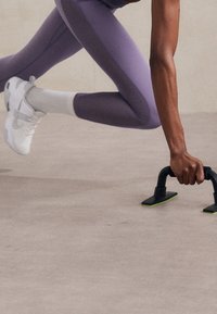 Fioletowe legginsy sportowe z wyraźnym wzorem szwów, zestawione z białymi trampkami. Osoba korzysta z czarnych i zielonych uchwytów do pompek na gładkiej powierzchni.