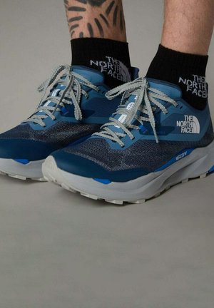 Baskets de course bleues avec une tige en mesh texturé, rehaussées par un logo blanc, des lacets gris et une semelle grise et bleue, portées avec des chaussettes noires.