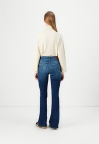 Krema pulover z visoko ovratnikom v kombinaciji s temno modrimi širokimi kavbojkami. Kavbojke imajo visok pas in dve zadnji žepki, s kožnim znakom na pasu.