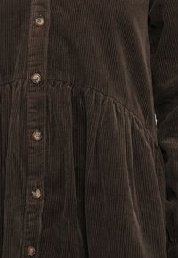 Indumento in corduroy marrone scuro con righe verticali, chiusura frontale con bottoni e cucitura arricciata in vita.