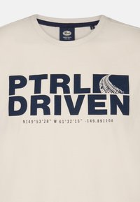 Krémové bavlněné tričko s výrazným námořnickým textem: "PTRLD DRIVEN" a geografickými souřadnicemi, se grafikou stop po pneumatikách.