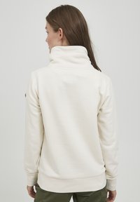 Lichte crèmekleurige sweatshirt met een opgerolde kraag, gemaakt van zacht materiaal; heeft een geribde onderkant en manchetten, met een patchdetail op de mouw.
