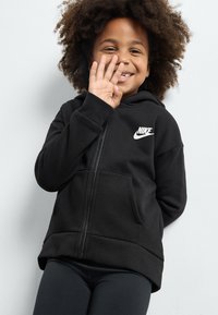 Svart zip-up hoodie i mjukt material, med en framficka och en vit Nike-logotyp på vänster bröst. Bärs med svarta leggings.