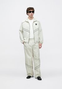 Ανοιχτό μπεζ φόρμα με μαύρες λεπτομέρειες, που περιλαμβάνει ζακέτα με φερμουάρ και παντελόνι με ελαστική μέση· συνδυάζεται με μαύρα γυαλιά ηλίο υ και μαύρα παπούτσια.