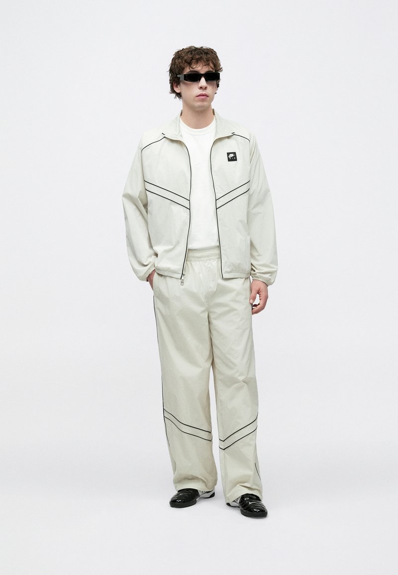 Chándal beige claro con detalles negros, que incluye una chaqueta con cremallera y pantalones con cintura elástica; combinado con gafas de sol oscuras y zapatos negros.