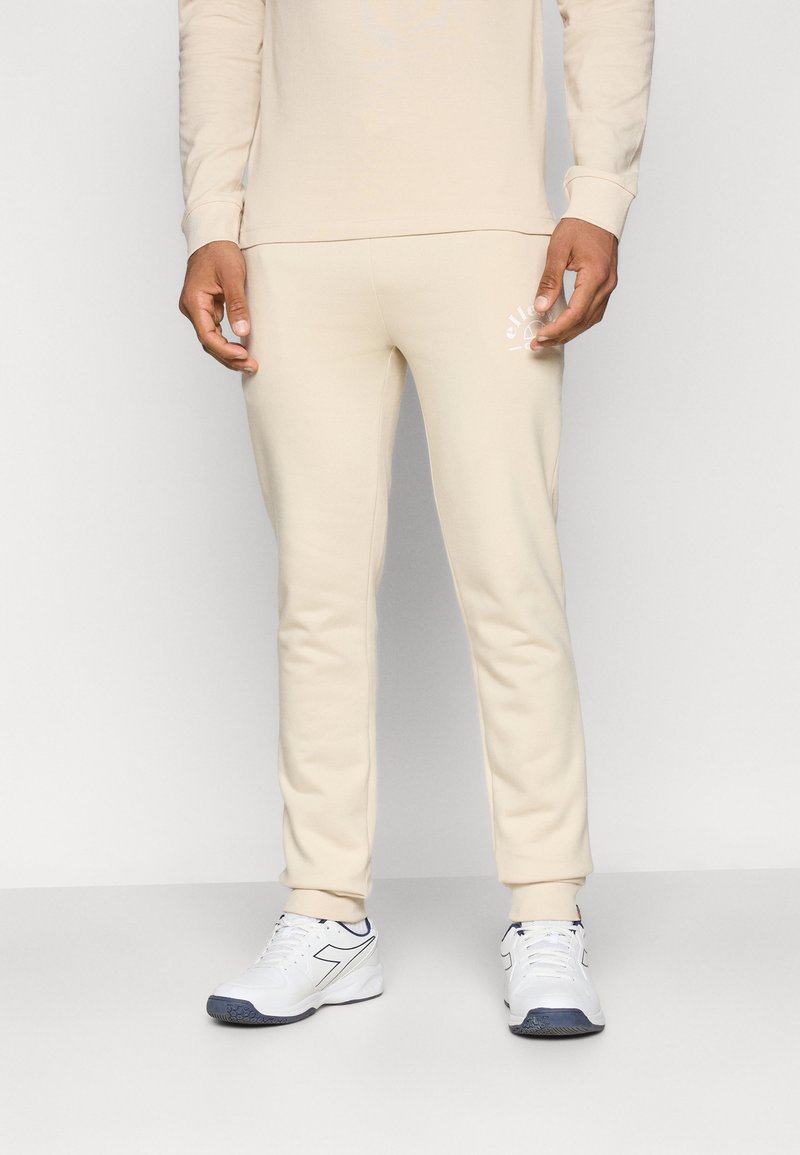 Mann in beigen Jogginghosen und passendem Oberteil mit weißen Turnschuhen, vor einem schlichten hellen Hintergrund stehend.