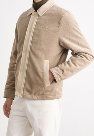 Mężczyzna w beżowej kurtce sztruksowej z zatrzaskami na oliwkowym golfie, ręka w kieszeni, zestawione z kremowymi spodniami na białym tle.