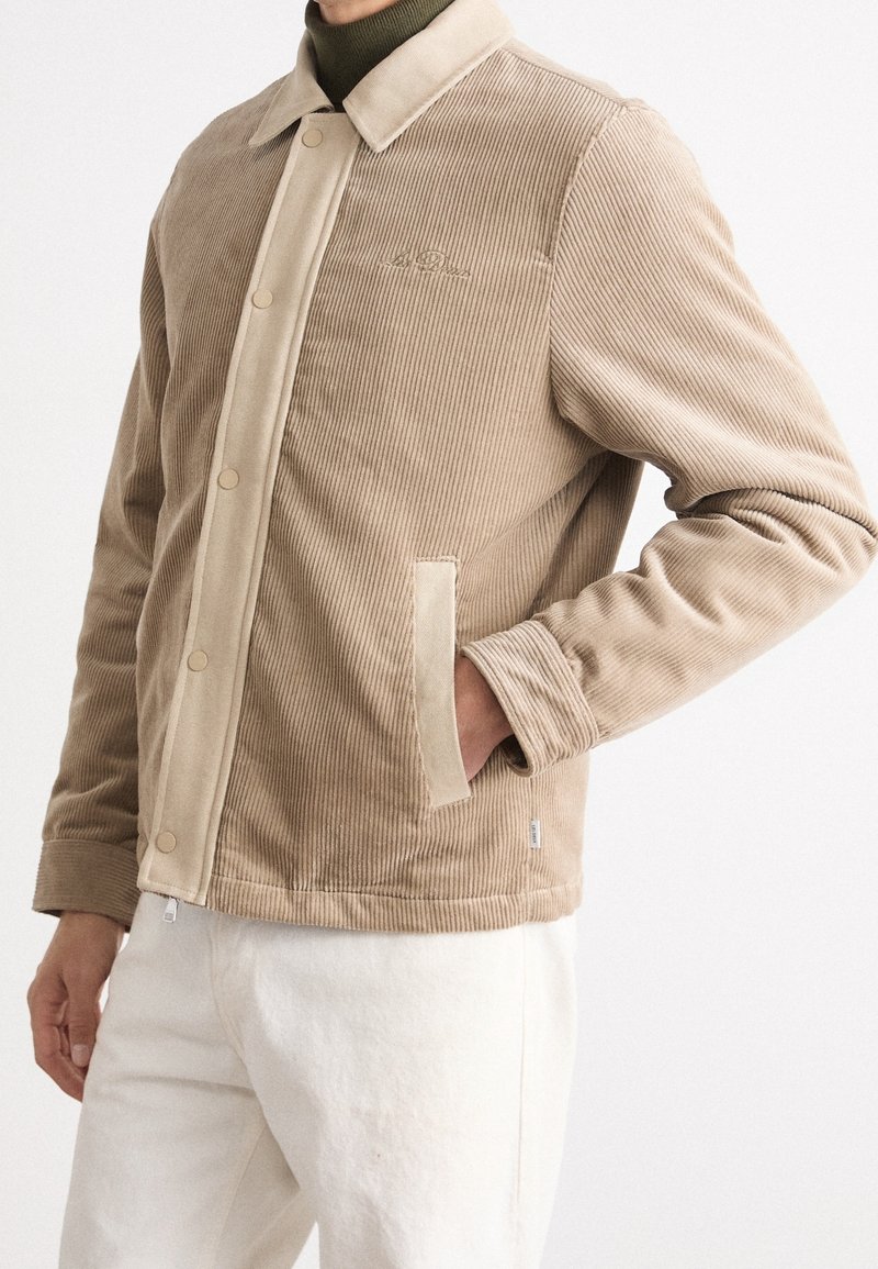 Homme portant une veste beige en velours côtelé avec des boutons-pression sur un col roulé olive, main dans la poche, associée à un pantalon écru sur fond blanc.