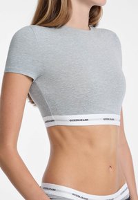 T-shirt court gris à manches courtes en tissu doux, doté d'une bande blanche avec le texte "GUESS JEANS" le long du bas. Texture lisse.