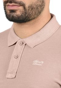 Lichtroze polo shirt van een textuurstof, met een placket met twee knopen en een klein wit logo geborduurd op de borst.