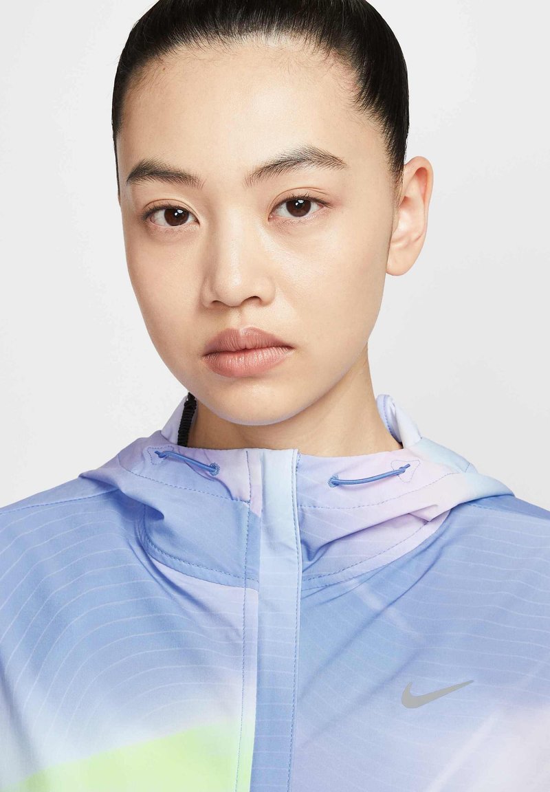 Lichtgewicht, pastelblau sportjack met een capuchon, ritssluiting aan de voorkant en subtiele gestreepte textuur. Heeft een klein zilveren Nike-logo op de borst.