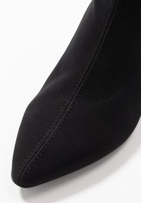 Bottine noire à bout pointu, fabriquée en tissu extensible, avec une texture lisse et des détails de couture visibles le long du côté.