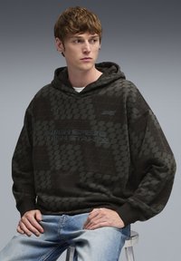 Felpa nera con cappuccio e motivo geometrico, caratterizzata da un logo e dal testo "HIGH SPEED. HIGH STAKES." Testo in blu, tessuto morbido, vestibilità rilassata, indossata con denim.