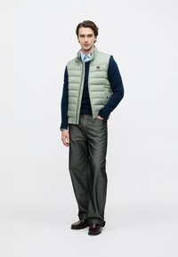 Light green puffer vest over a navy sweater, gray pleated trousers, and dark shoes. Features a logo on the vest and textured fabric.