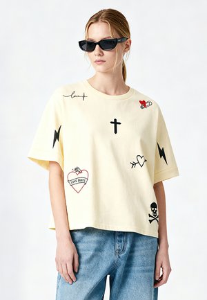 Femme portant un t-shirt beige avec des imprimés graphiques noirs et rouges comprenant des cœurs, une croix, des éclairs, un crâne et du texte, associé à un jean bleu et des lunettes de soleil.
