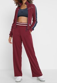 Bordeauxfarbener Sportanzug mit weißen und marineblauen Akzenten; Jacke mit Reißverschluss und weit geschnittene Hose mit Knopfdetails am Saum; glattes Material.