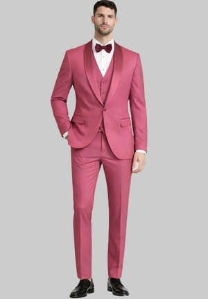Homme portant un costume trois pièces rose ajusté avec un revers châle en satin, une chemise blanche, un nœud papillon bordeaux et des chaussures habillées noires, debout devant un fond gris.