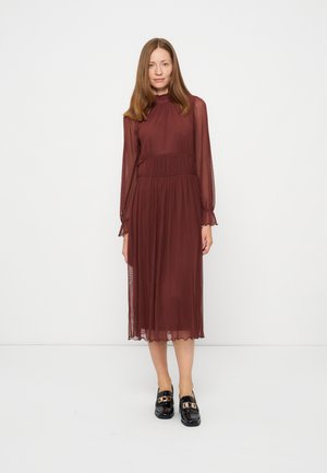 Femme aux cheveux bruns raides portant une robe midi bordeaux à manches longues et des mocassins noirs ornés de détails en chaîne dorée, debout sur un fond blanc uni.