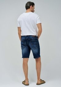 Pantalones cortos de mezclilla azul con un ajuste regular, diseño de cinco bolsillos, ligero desgaste en la parte delantera y un parche de cuero en la cintura trasera. Camiseta blanca.