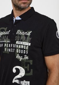 Melnā polo krekls, kas izgatavots no kokvilnas maisījuma, ar apkakles kaklu, ar dažādas tipogrāfijas grafiku baltā un zaļā krāsā, un logotipa uzšuvumu uz piedurknes.