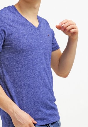 Man draagt een blauw T-shirt met V-hals en korte mouwen met een klein logo op de borst, en past met zijn rechterhand de mouw aan tegen een effen achtergrond.