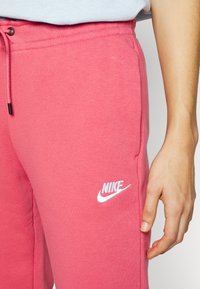 Růžové bavlněné tepláky s elastickým pasem a stahovací šňůrkou, s bílým logem Nike na přední straně a bočními kapsami.