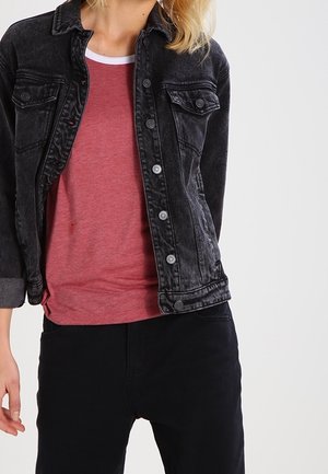 Vrouw die een vervaagde zwarte spijkerjas draagt over een rood overhemd met witte bies en een zwarte broek, staand tegen een eenvoudige lichte achtergrond.