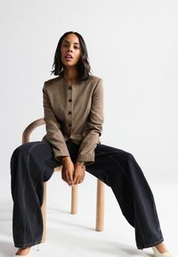 Brown houndstooth jacket with round neckline and black buttons, paired with wide black jeans. Sitting on a wooden chair, neutral background.