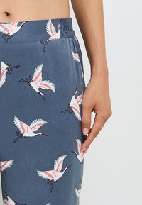 Pantalons bleus à mi-taille avec une ceinture élastique, arborant un motif de grues répété en rose et blanc sur un tissu lisse.