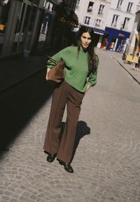 Femme en pull vert et pantalon marron marchant dans une rue pavée, tenant un sac marron dans un milieu urbain.