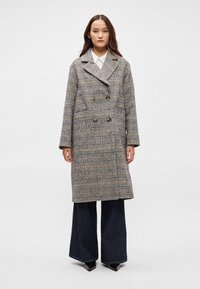 Διπλόχρωμο καρό παλτό σε γκρι, μπεζ και μαύρο. Ξεχωρίζει για το υπερμεγέθη σχήμα του, το μεγάλο γιακά και τα δύο κουμπιά στο μπροστινό μέρος.