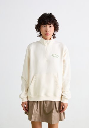 Crème-kleurige sweatshirt met kwartrits en een voorkantzak, met geborduurd "New Balance Athletics" in het groen. Zachte, gladde textuur.
