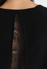 Schwarzes Stricktop mit einer schmalen Rückenöffnung, die aufwendige schwarze Spitze mit floralem Muster darunter sichtbar macht.