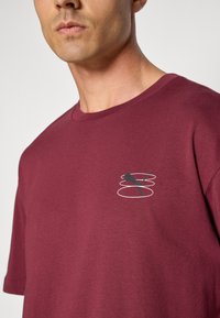 Bordeaux bomulds T-shirt med et lille grafisk design af en springende figur omkranset af tre hvide ringe på venstre brystområde.
