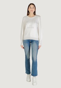 Witte gebreide sweater met "GUESS" logo, zilveren accenten, lange mouwen; gecombineerd met lichtblauwe spijkerbroek en witte sneakers.