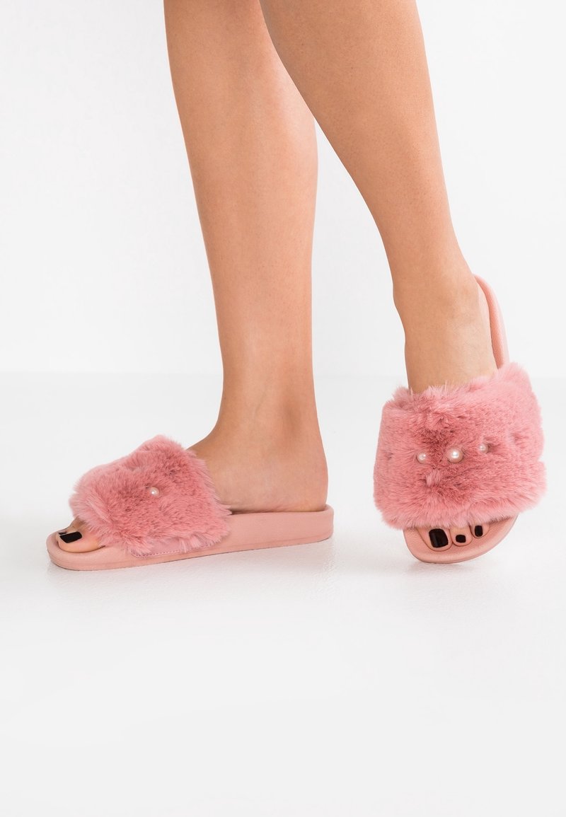 Pieds portant des sandales à enfiler roses et duveteuses ornées de petites perles décoratives sur les brides, avec un vernis à ongles noir visible sur les orteils.