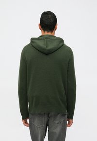 Zöld kapucnis pulóver puha anyagból, nagy kapucnival, hosszú ujjakkal és laza szabással, szürke farmernadrággal párosítva.