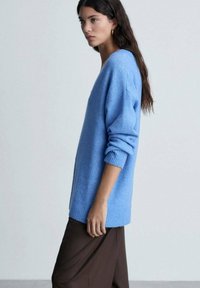 Maglione a maglia di colore azzurro chiaro con scollo a V e polsini a coste, indossato su pantaloni marroni larghi. Texture liscia, design casual.