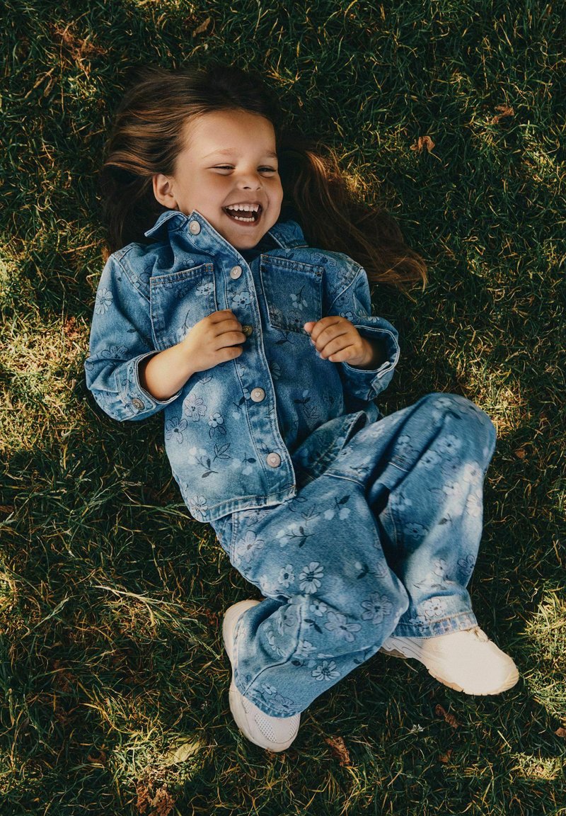 Jeune fille riant et allongée sur l'herbe, portant une veste en jean bleue à fleurs et un pantalon avec des baskets blanches.