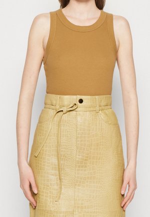 Donna che indossa un top marrone senza maniche e una gonna beige a vita alta con texture effetto coccodrillo e cintura annodata, in piedi su sfondo bianco.