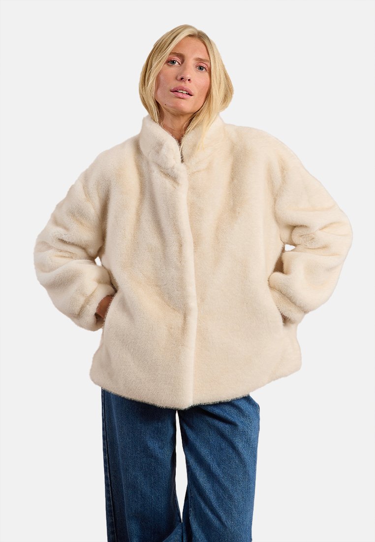 Giacca in pelliccia finta di colore crema con colletto alto, vestibilità oversize, spalle drop e maniche larghe. Abbinata a jeans a gamba larga blu.