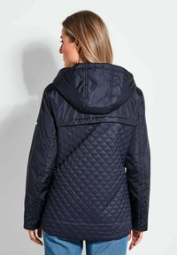 Marinegeklede gewatteerde jas met capuchon, voorzien van een diagonaal gestikt patroon en gladde schouderpanelen, getoond van de achterkant.