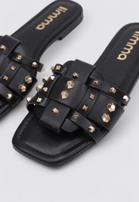Des sandales à glisser en faux cuir noir comportent une large bride ornée de clous dorés de différentes formes, et un design à bout carré.