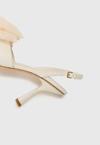 Hvite hæler med tynne stropper, en støttende ankelspenne og en teksturert nylon sløyfedetalj ved tåen. Slank, stiletto hældesign.