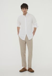 Camicia bianca con colletto alla cinese, pantaloni beige a righe verticali, e scarpe slip-on in suede marrone. Tessuto liscio, vestibilità rilassata.
