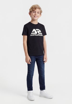 Lyshåret dreng iført sort DSquared2-logo t-shirt, blå jeans og hvide sneakers, stående mod en ensfarvet hvid baggrund.