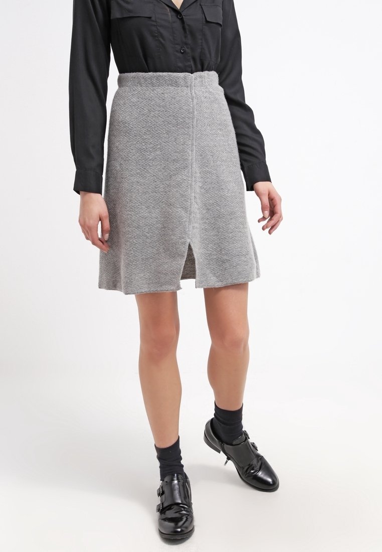 Jupe grise texturée avec un design évasé et une fente sur le devant. Associée à un chemisier noir à boutons et des chaussures noires brillantes.