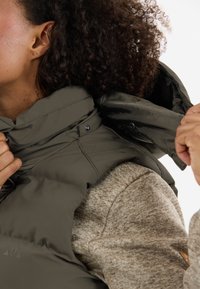 Dunkelgrüne isolierte Weste mit hohem Kragen und Druckknöpfen, über einem strukturierten beigen Pullover geschichtet. Fokus auf Funktionalität und Wärme.