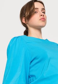 Top azul brilhante feito de um tecido suave. Apresenta um decote redondo e mangas bufantes. O design é simples, sem padrões ou acentos visíveis.